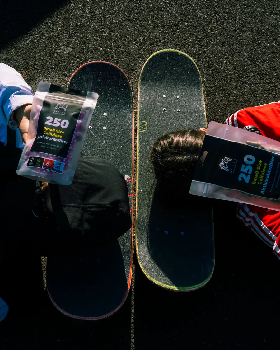 Zwei Skateboards auf Asphalt, daneben halten zwei Leute je eine 250er-Packung Kailar Filter hoch
