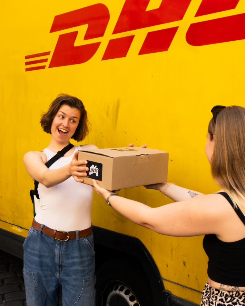 Zwei Frauen reichen sich vor einem gelben Lieferwagen ein Kailar-Paket